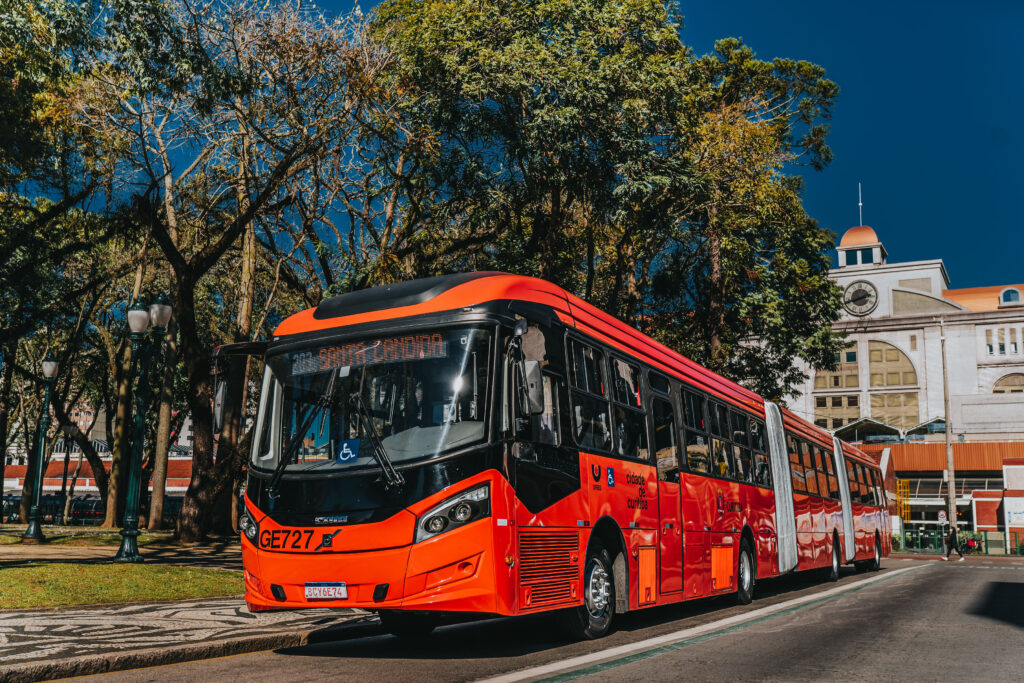 Um ônibus biarticulado é visto circulando ao lado de uma praça de Curitiba