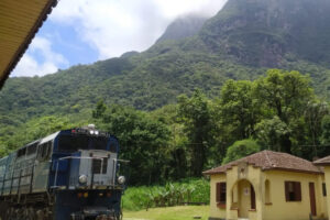 Trem passa pela estação Marumbi no pé do pico Marumbi. O trem está em primeiro plano, a câmera está apontada ligeiramente para cima. O pico Marumbi está em último plano.