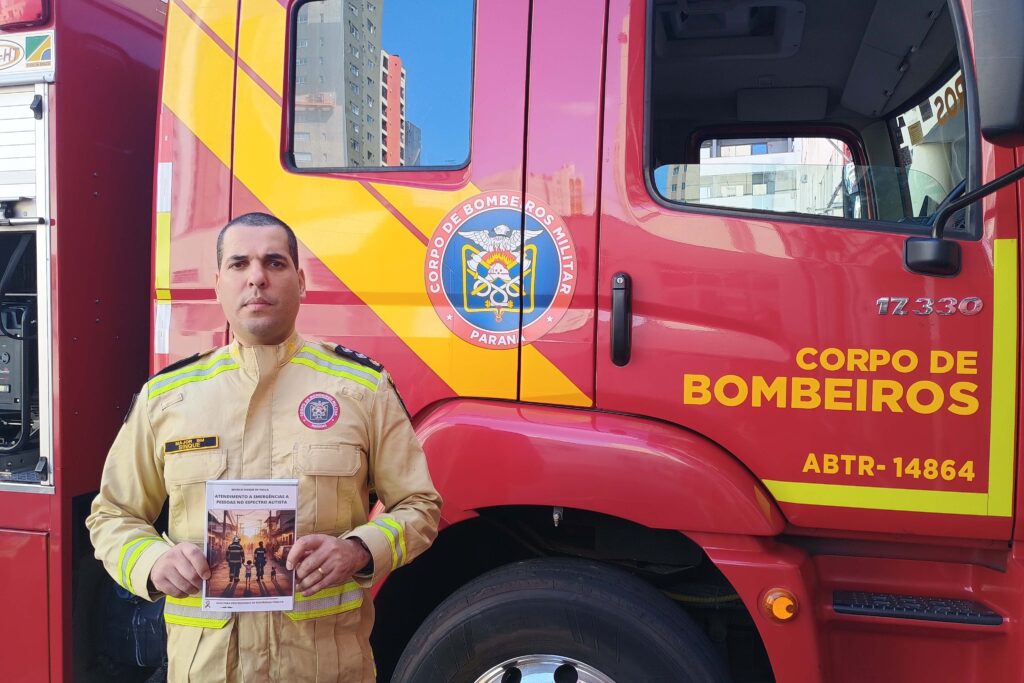 Major Murilo Sinque de Paula posa com o manual em mãos ao lado de caminhão dos bombeiros.