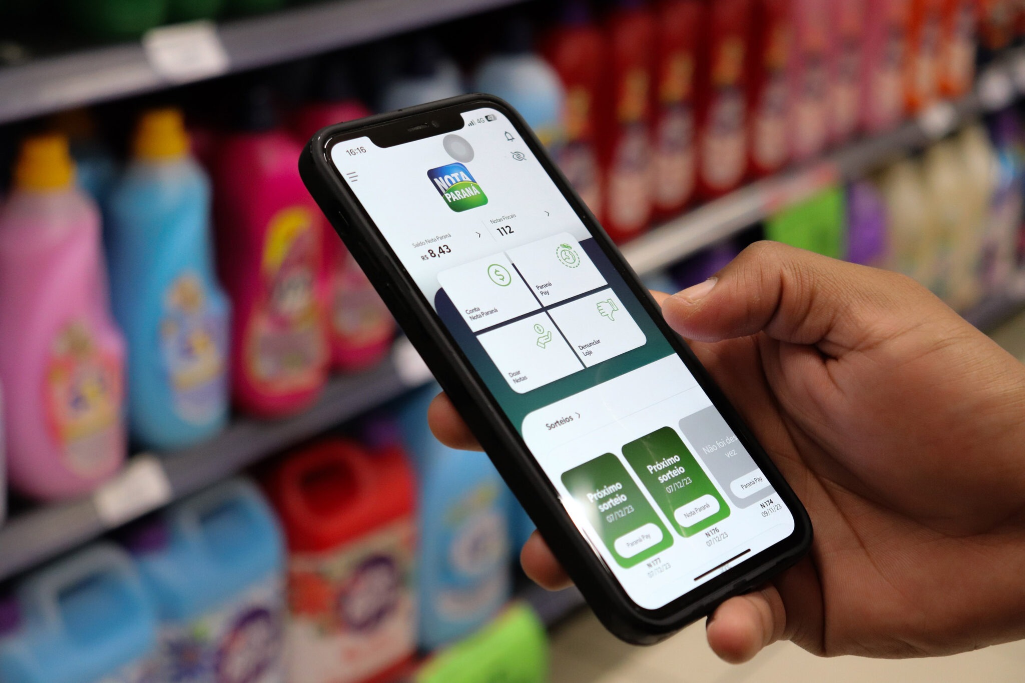 Pessoa segura celular com aplicativo da Nota Paraná em frente a prateleira de mercado