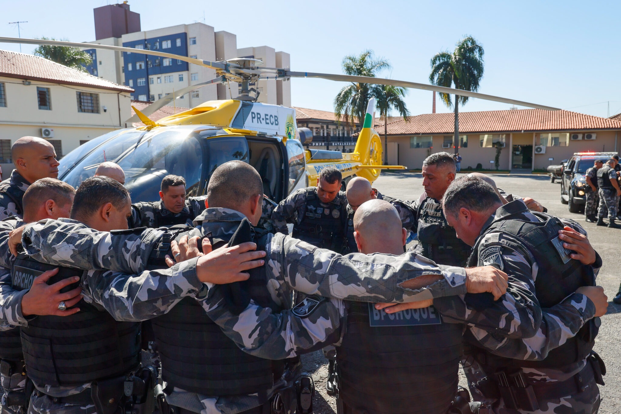 Policiais Militares se abraçam em círculo. Ao fundo, um helicóptero pousado.