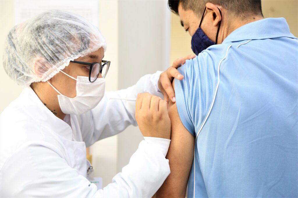 Profissional de saúde em uniforme aplica vacina em braço de paciente. Ambos usam máscaras