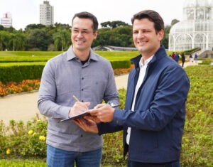 Deltan Dallagnol segura "Carta de Compromisso com Curitiba" enquanto Eduardo Pimentel assina. Ambos sorriem para câmera