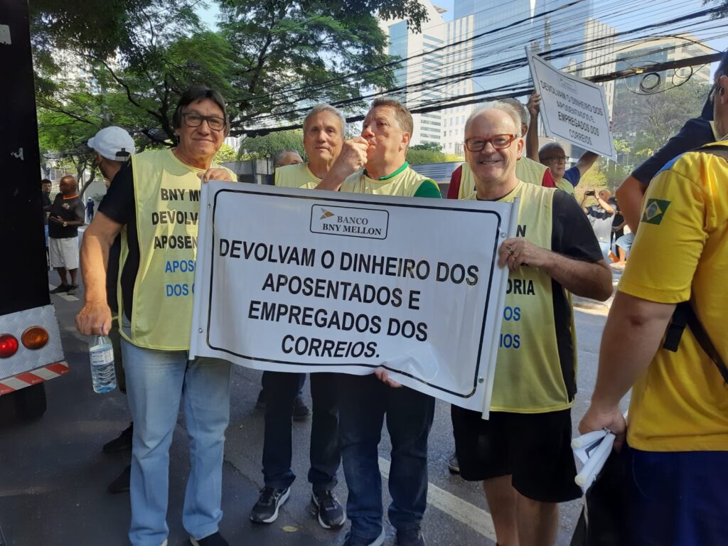 Membros dos correios seguram faixa escrita "Devolvam o dinheiro dos aposentados e empregados dos correios". Todos estão vestindo coletes amarelos.