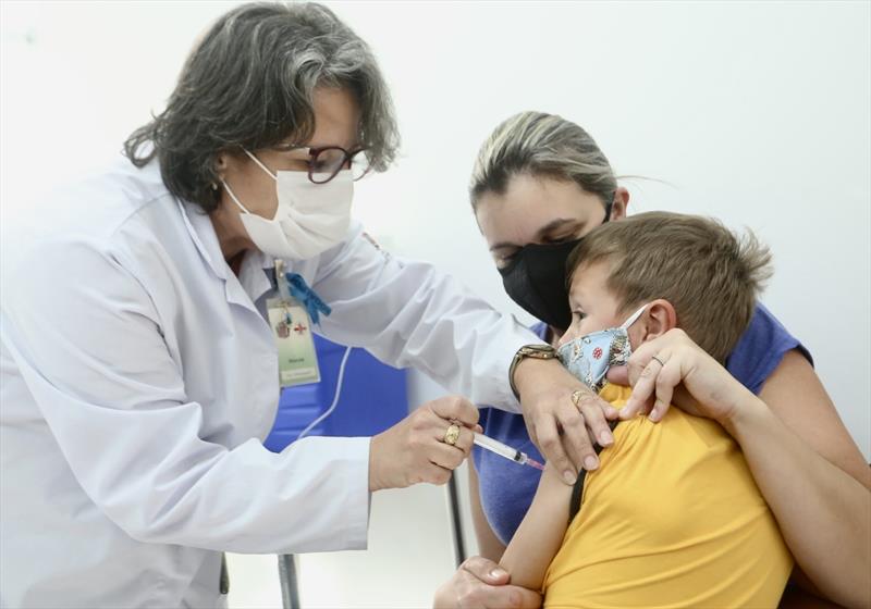Enfermeira aplica vacina em criança no colo de mãe.