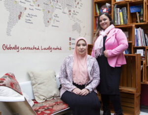 Duas mulheres lado a lado. Uma, vestindo hijab, está sentada num sofá. A outra, em pé, logo atrás do ombro da primeira