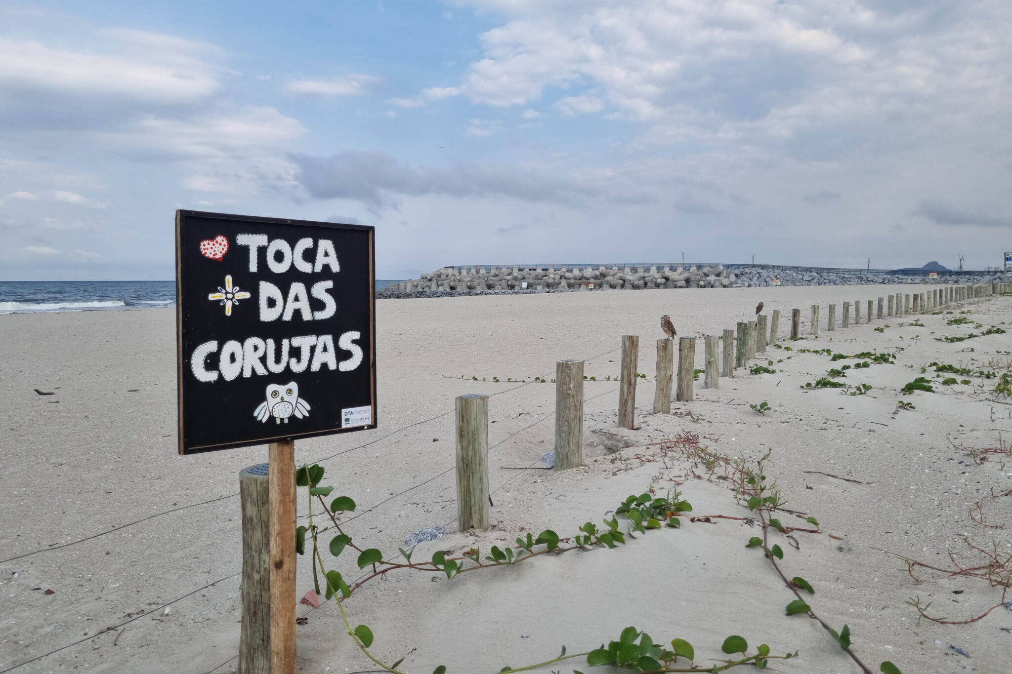 Cerca separando a praia da área de restinga. Uma placa de fundo preto e escrita branca tem escrito: "Símbolo de coração, toca das corujas símbolo de coruja"