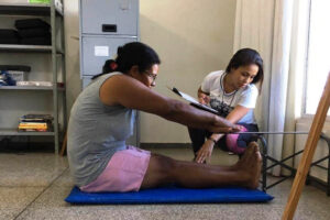 Pessoa sentada sob tapete faz teste de mobilidade enquanto profissional de educação física faz a medição do teste