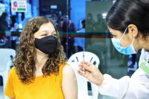 Paciente com camiseta amarela e máscara de pano preta inspeciona vacina na mão de profissional da saúde que veste um jaleco branco e uma máscara descartável azul.