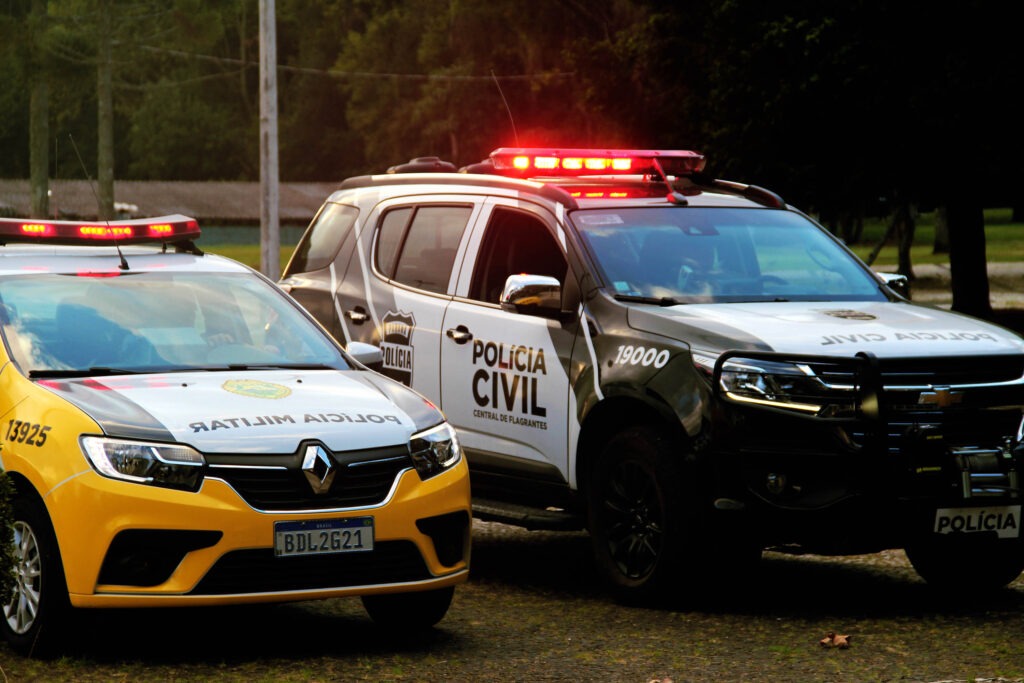 Duas viaturas lado a lado. A viatura da esquerda é amarela e branca, pertence a Polícia Militar. A viatura da direita, é preta e branca e pertence a Polícia Civil.