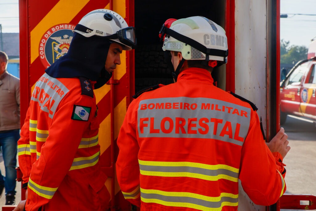 Dois bombeiros de costas. Na parte de cima das costas da jaqueta está escrito: Bombeiro Militar Florestal