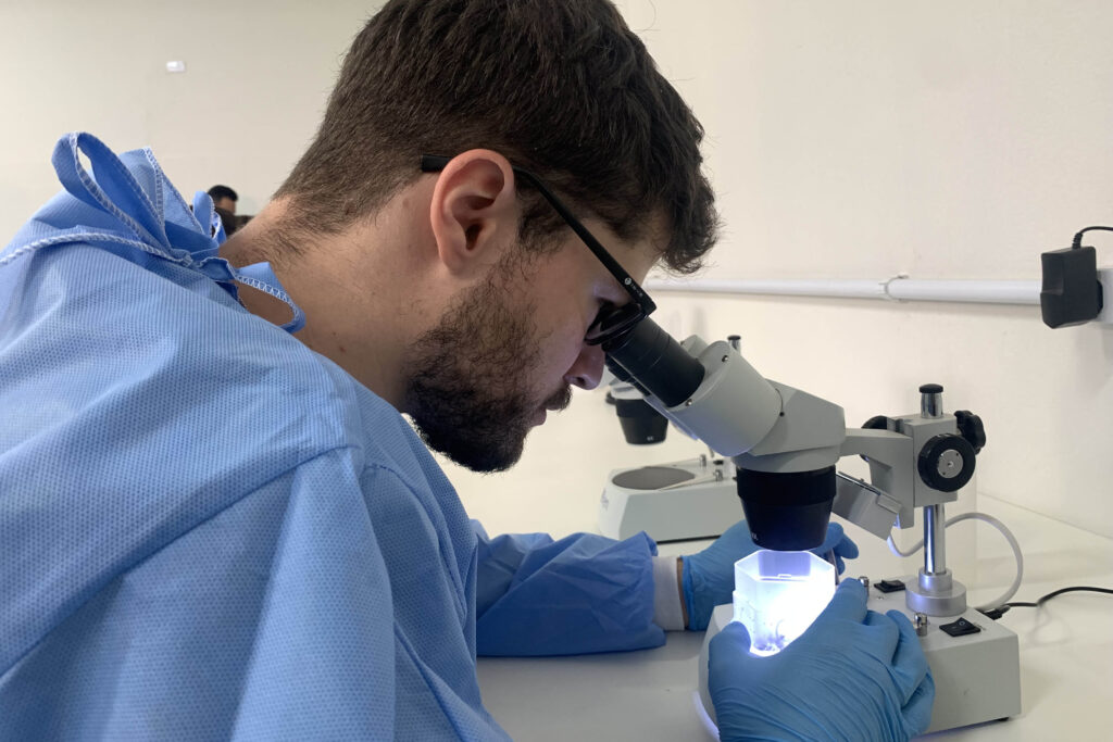 Pessoa vestindo uniforme de laboratório observa frasco através de microscópio.