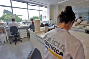 Interior de laboratório da Polícia Científica. Em primeiro plano, uma pessoa vestindo jaleco branco está de costas para a câmera. Nas costas do jaleco branco está escrito em preto e cinza "Polícia Científica"