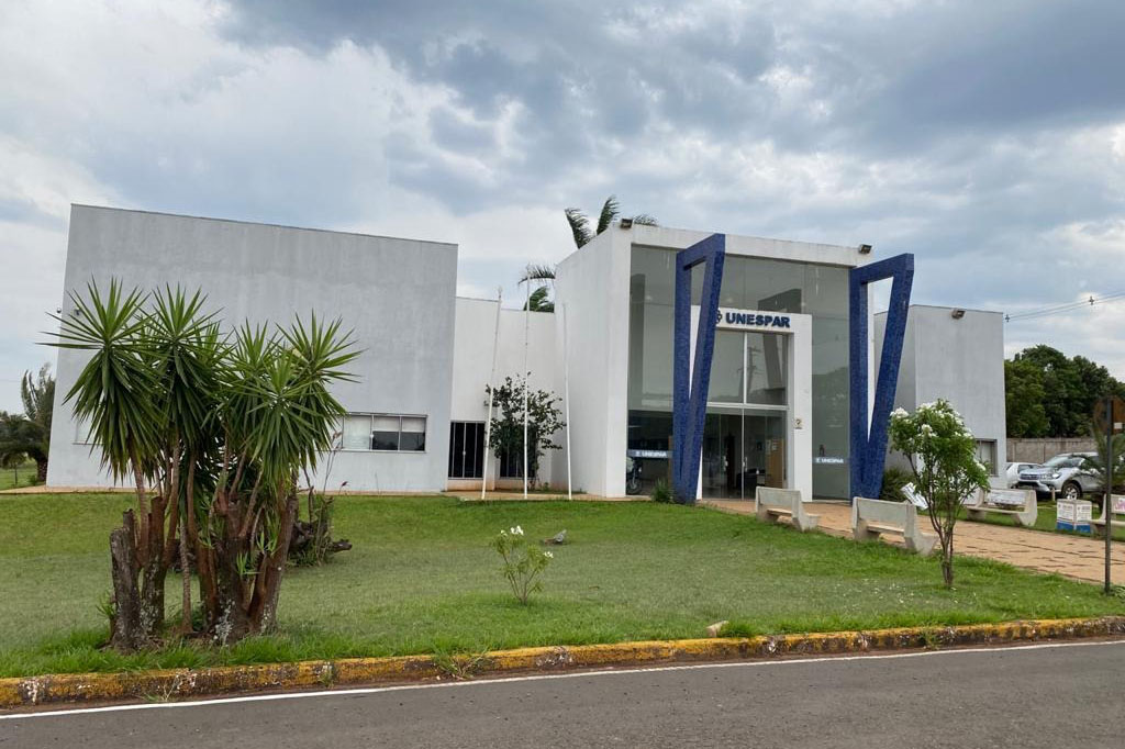 Fachada da UNESPAR. Em primeiro plano está parte do asfalto, o meio fio e uma faixa de grama. O prédio na beira da grama é branco com colunas azuis.