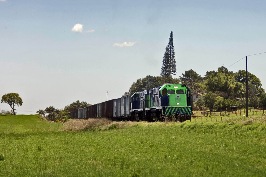 Trem com diversos vagões vem da esquerda para a direita. A cor dos vagões é verde e tem grama no primeiro plano