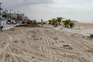 Praia de Matinhos com funcionários uniformizados com coletes refletores e maquinário pesado como caminhões de carga