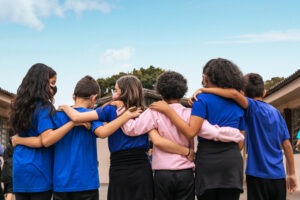 Seis crianças de costas para a câmera unem os braços em corrente. Eles vestem uniforme azul marinho e um deles veste moletom rosa claro