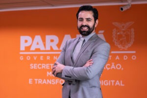 Rafael Sanchez Loureiro posa em frente a parede de cor laranja. Na parede está o logotipo do governo do estado e em letras brancas e caixa alta está escrito: Secretaria da Inovação, Modernização e Transformação Digital. Loureiro está vestindo um terno cinza e uma gravata cinza claro. Ele tem barba e cabelo escuros e pele branca.