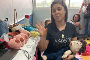 Mulher com camiseta "Agosto Dourado" faz demonstração com boneco de tricô