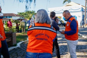 Técnicos da Defesa Civil, vestindo coletes laranja, conversam com moradores de Morretes sobre o teste do alerta climático.