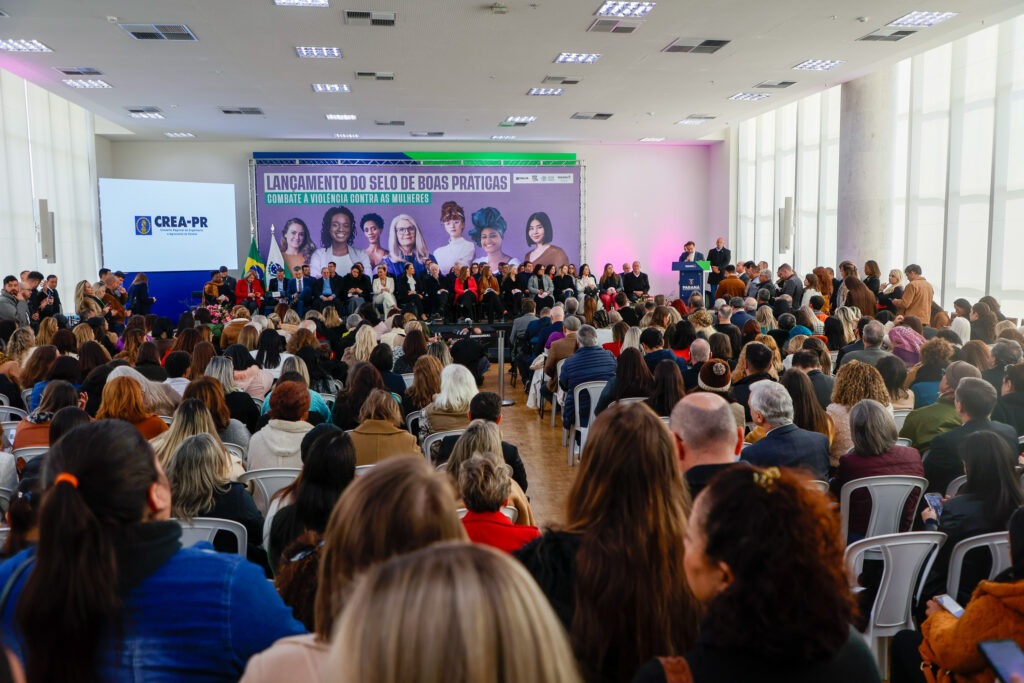 Salão com pé direito alto cheio de pessoas, existe apenas um corredor estreito entre as pessoas sentadas. Ao fundo do salão, um palco com um banner retangular que vai de ponta a ponta do palco e até o teto. No banner está escrito "Lançamento do selo de boas práticas"