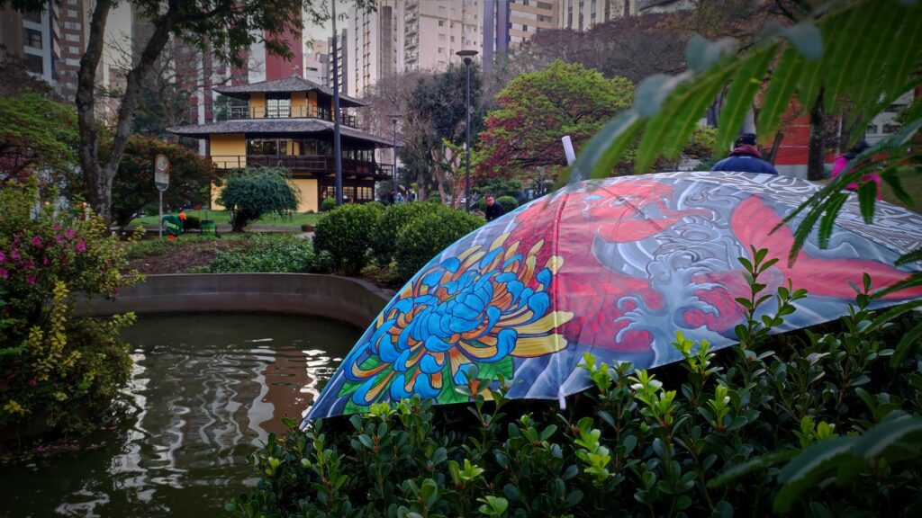 Guarda-chuva com arte em primeiro plano. Em segundo plano, uma pequena lagoa e um prédio baixo com estilo japonês.