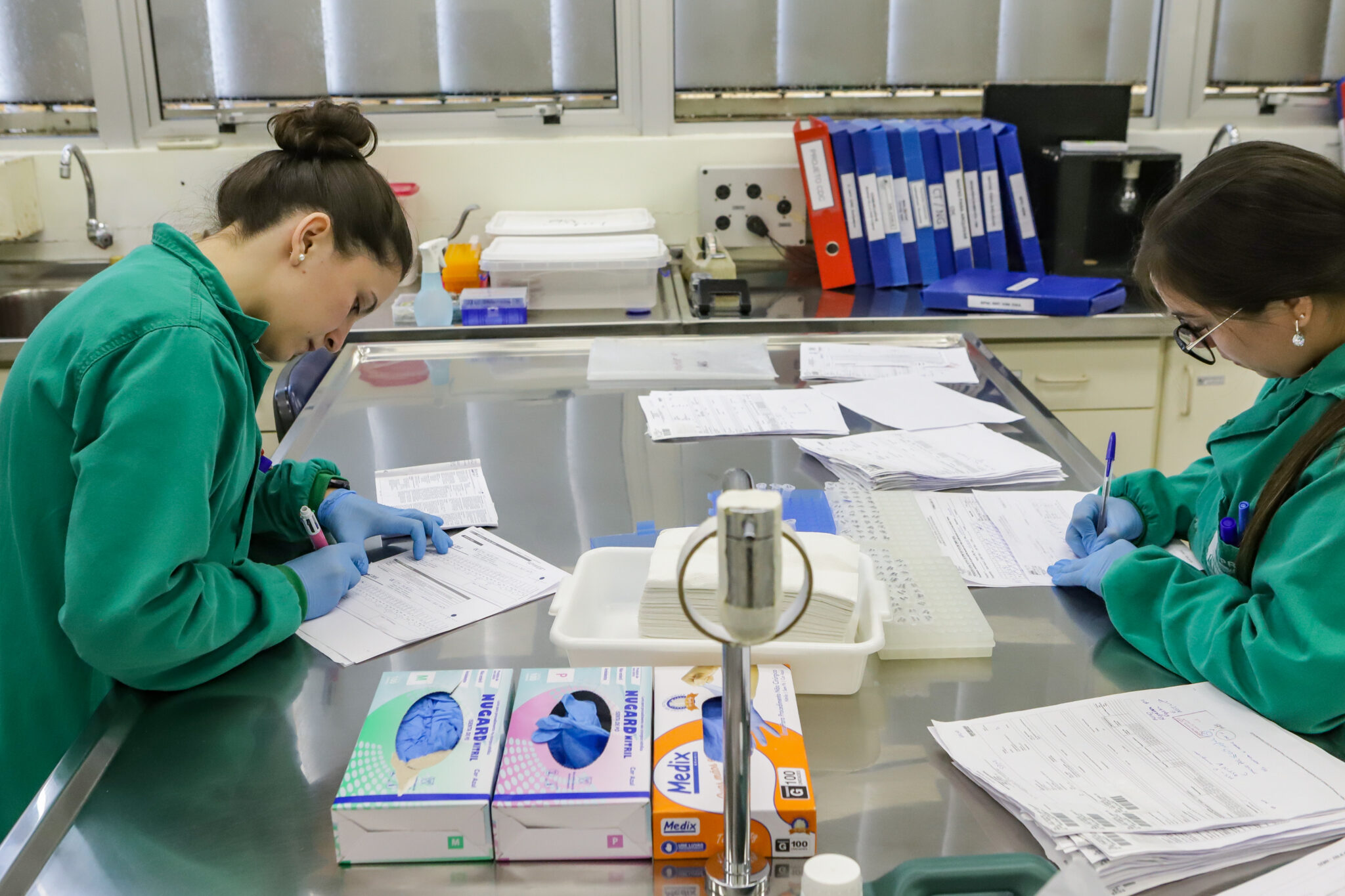 Duas pessoas preenchem formulários em laboratório. Ambas as pessoas estão vestindo roupas verdes de algodão.