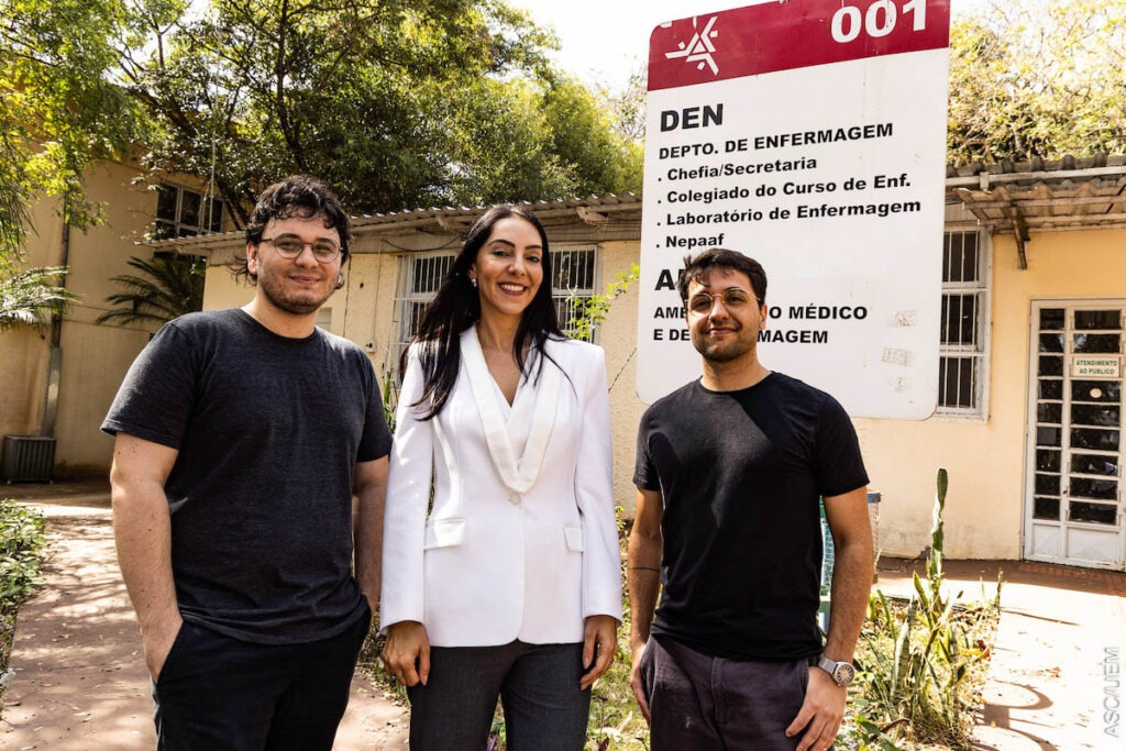 Da esquerda para a direita, Lucas Vinícius de Lima, Gabriela Magnabosco e Gabriel Pavinati, pesquisadores integrantes do Gepvhat/UEM. Três pessoas posam em frente à placa com direções. As duas pessoas às pontas têm apresentação masculina e vestem camisetas pretas, a pessoa ao meio tem apresentação feminina e veste um paletó branco.