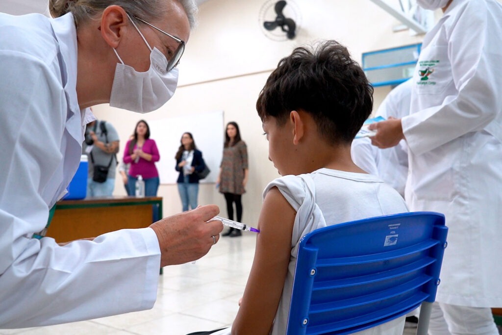 Criança sentada em cadeira de costas para a câmera, recebe vacina no braço esquerdo de profissional de saúde vestindo máscara hospitalar e jaleco branco.