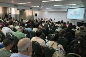 Pessoas em auditório observam apresentação de powerpoint sendo projetada ao lado de mesa com pessoas viradas à plateia