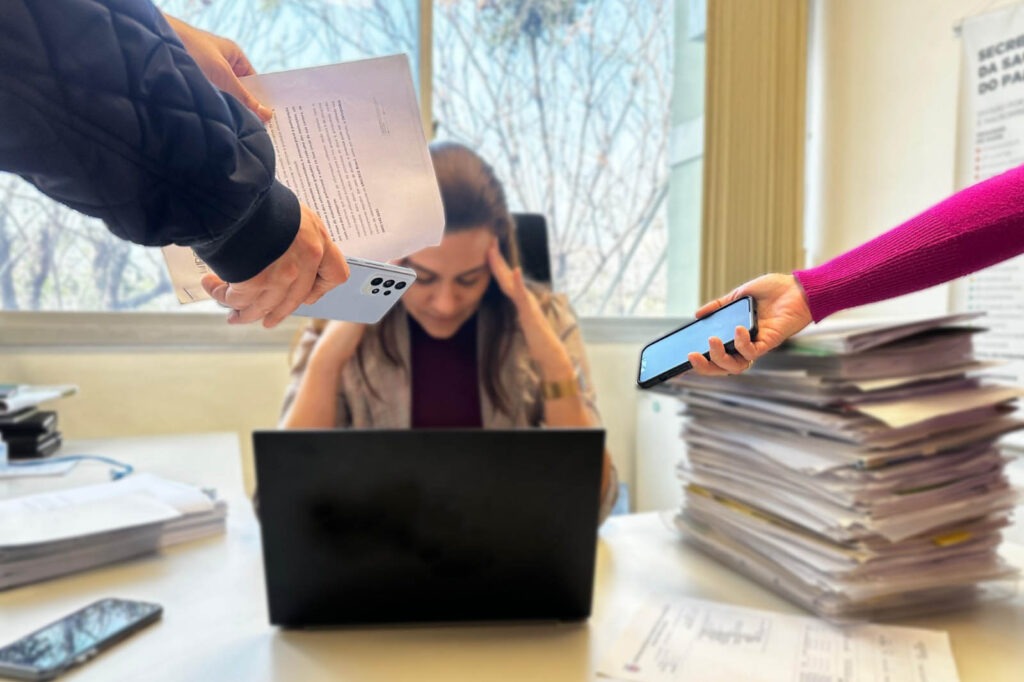 Uma pessoa atrás de um computador segura sua cabeça com as duas mãos. Mãos vindo de ambos os lados da tela tentam chamar a atenção da pessoa com celulares e papéis.