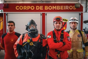 Quatro bombeiros posam em frente à caminhão de bombeiros. Da esquerda à direita: uniforme de mergulho sem capacete, uniforme de mergulho em profundidade com tanque de oxigênio e máscara, uniforme leve para rapel e uniforme leve para combate à chamas sem máscara.