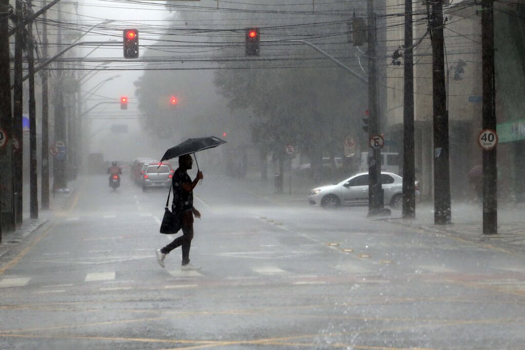 Pessoa atravessa a rua da esquerda para a direita. A pessoa carrega um guarda-chuva e ao fundo podemos ver um semáforo ofuscado pela chuva.