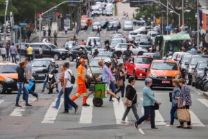 Pessoas atravessam a faixa de pedestres em primeira mão. Ao fundo, vários carros