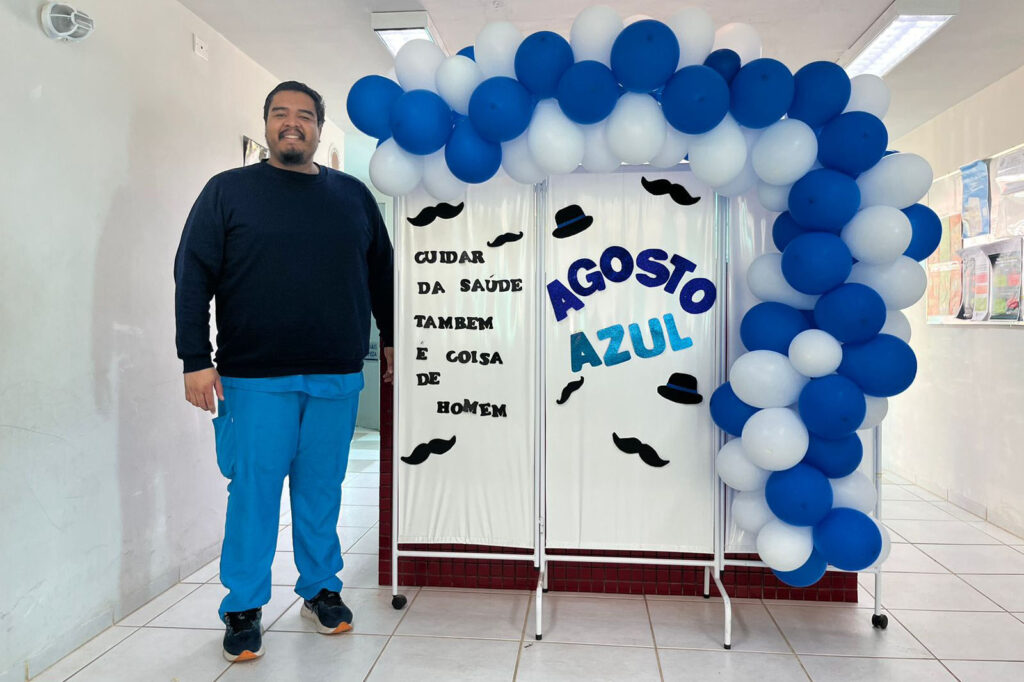 Homem posa ao lado de painel onde está escrito: "Agosto Azul. Cuidar da saúde também é coisa de homem". O painel está decorado com balões azuis e brancos.