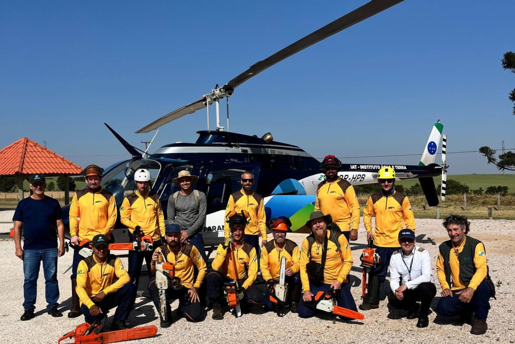 Técnicos e voluntários posam em frente a helicóptero. Eles estão uniformizados de amarelo e alguns vestem capacetes