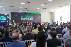 Salão de paredes e teto brancos. Em primeiro plano, várias filas de cadeiras apontadas para frente. Ao fundo do salão, um palco com diversas pessoas sentadas na direção da plateia. No fundo do palco, tapando a parede, um painel escrito "De homem para Homem" em letras verdes e fundo azul escuro. Uma mulher discursa num púlpito no lado esquerdo do palco