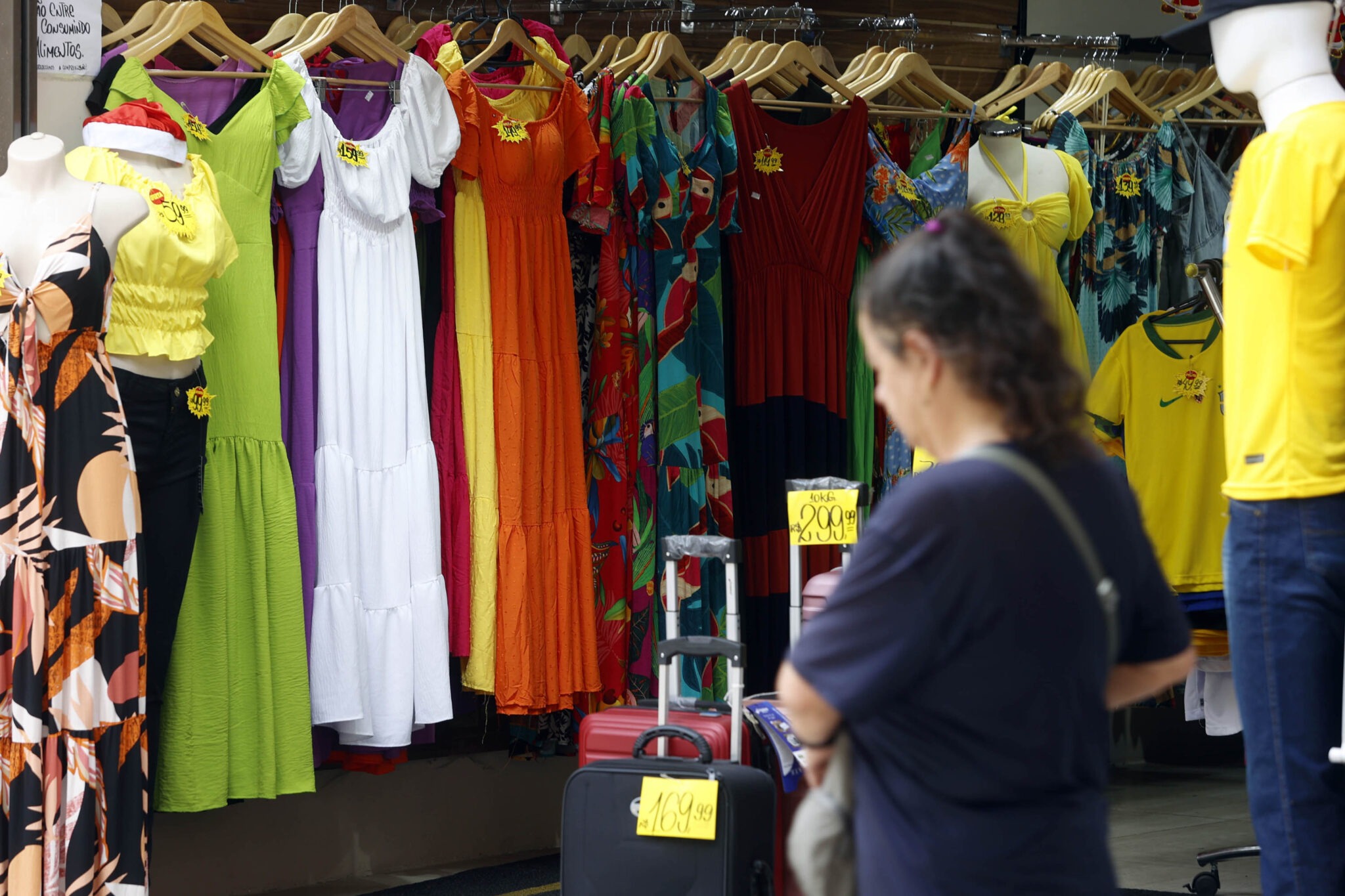Em primeiro plano, uma pessoa em desfoque caminha do lado direito para o esquerdo da foto. Ao fundo, em foco, vários vestidos pendurados em cabides estão dentro de uma loja.