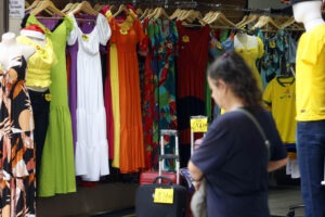 Em primeiro plano, uma pessoa em desfoque caminha do lado direito para o esquerdo da foto. Ao fundo, em foco, vários vestidos pendurados em cabides estão dentro de uma loja.