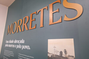 Painel de cor cinza. Em letras douradas e grandes Morretes. Abaixo do nome da cidade, em letra bold e branca, está escrito Parte da nossa história, Parte do nosso povo.