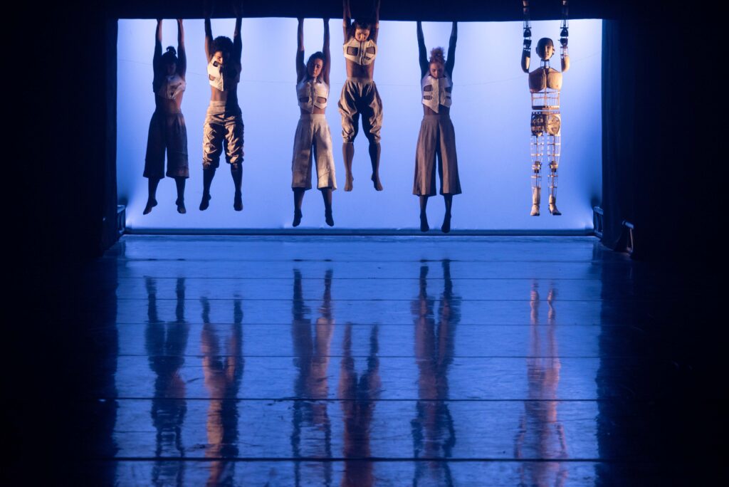 Bailarinos se penduram em estrutura no palco. Ao fundo, uma tela azul ilumina a silhueta dos bailarinos