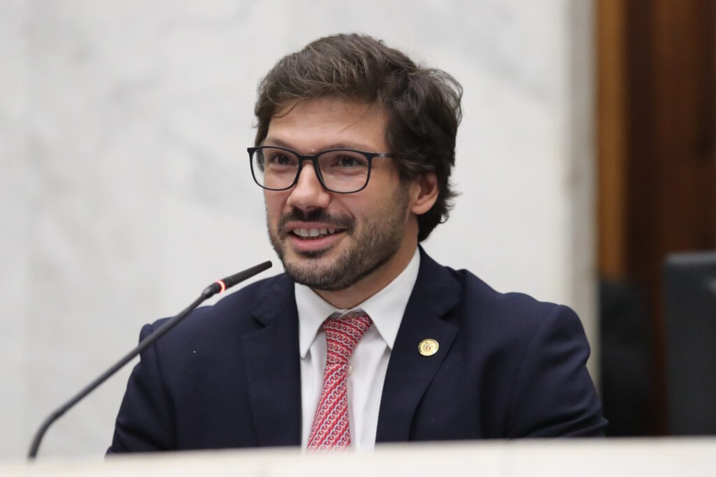 A imagem mostra Tiago Amaral, um homem branco, com cabelos curtos e castanho, barba e bigode, que usa óculos de armação em acetato escuro e quadrado. Ele aparece do peito para cima. Está atrás de um pulpito com microfone e usa terno e gravata.