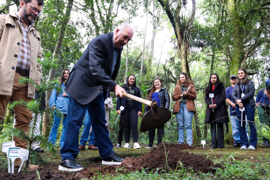 A câmera está quase encostando no chão, mirada de baixo para cima. Em primeiro plano, vemos uma pessoa de calça jeans, blazer usando uma enxada contra o chão. Em segundo plano, um semi-círculo de pessoas aplaude.