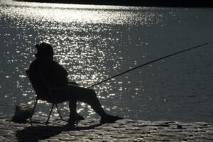 Pessoa pesca enquanto senta em cadeira. O reflexo da água ao fundo causa um efeito de backlighting na pessoa em primeiro plano, deixando a silhueta da pessoa totalmente preta mas perfeitamente delimitada em contraste com a água ao fundo