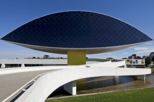 Fachada do Museu Oscar Niemeyer. Imagine uma estrutura elevada, suspensa por uma base amarela de concreto. A parte superior lembra o contorno de um olho gigante, alongado, com as extremidades afinando para os lados. Esse “olho” parece flutuar no ar, em cima de um pilar largo e maciço, o que dá a sensação de leveza, mesmo com sua grandeza. Ao redor do museu, há um espelho d'água, refletindo a construção e amplificando sua presença. As superfícies são lisas, com poucas texturas, predominando o concreto branco, criando um contraste marcante com o verde do entorno.