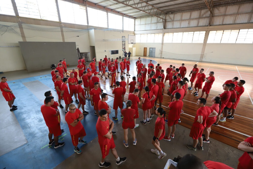 Bombeiros em uniforme de exercício se reúnem em quadra poliesportiva.