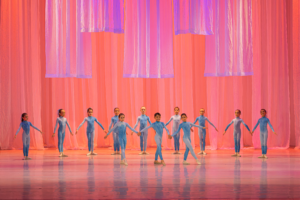 Crianças usando roupas azuis se apresentam em palco. Ao fundo, uma cortina rosa e vermelha.