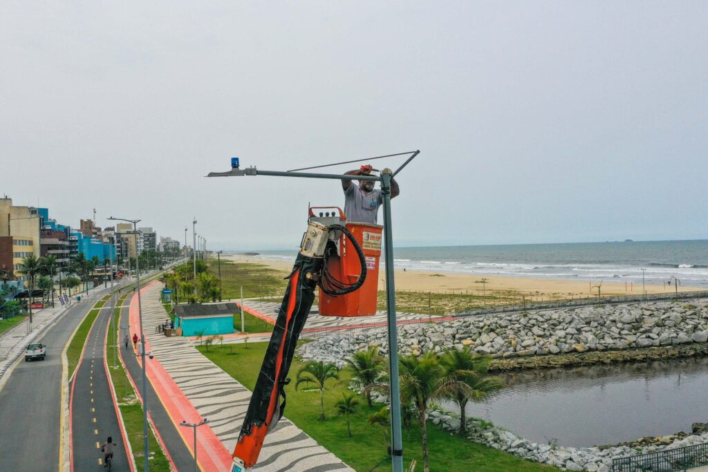 Operador usa de guindaste para alcançar topo de poste. Ao fundo, orla da praia de Matinhos