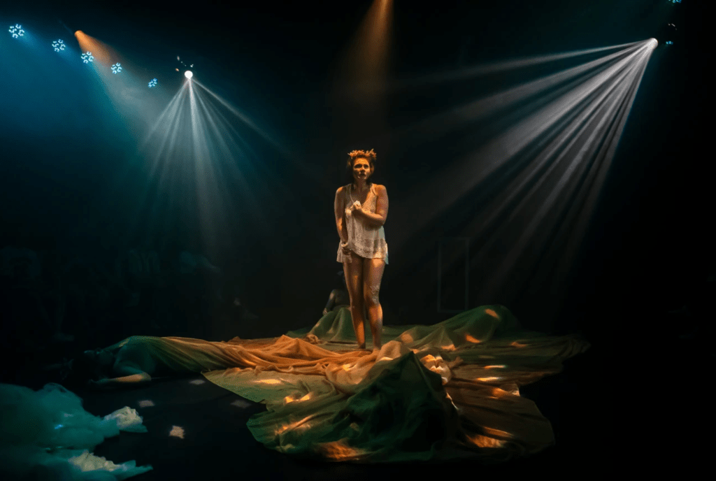 Uma pessoa no centro do palco, ela parece tentar se cobrir. Ao chão, panos em volta da personagem. Feixes de luz são apontados para a personagem, centralizando-a ainda mais