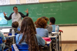 Sala de aula. Em primeiro plano, alunos sentados em carteiras de costas para a câmera. Ao fundo, um professor em pé e quadro negro